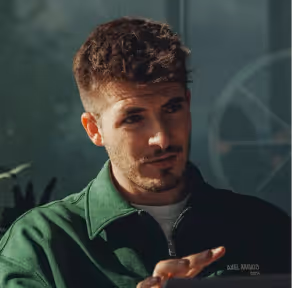 Young man with curly hair and beard wearing a green jacket, gesturing with his hand.