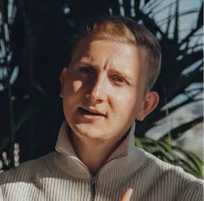 Man with short blonde hair wearing a light zip-up jacket, with green foliage and sunlight in the background.