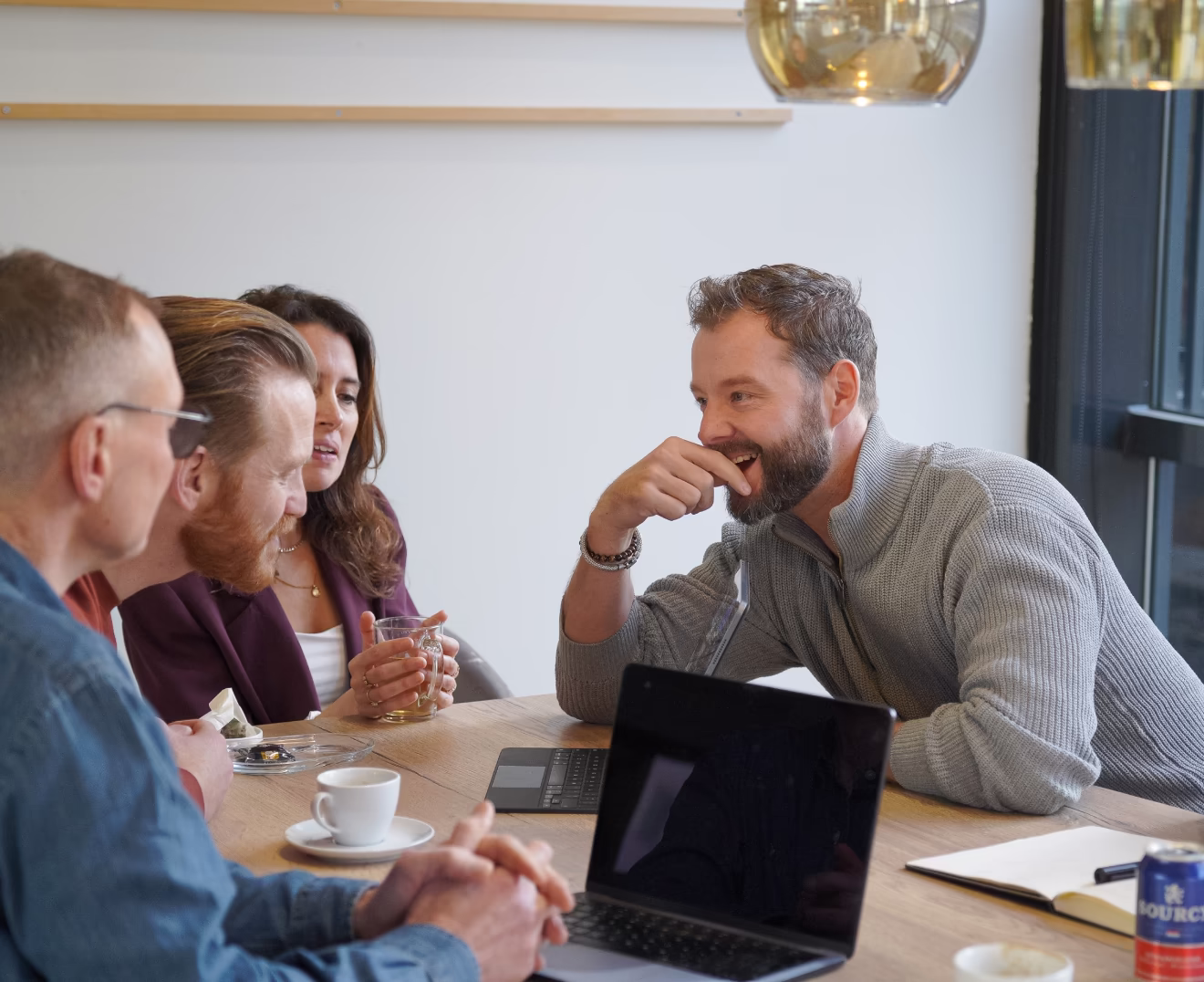 Vier volwassenen zitten aan een tafel in een lichte kamer, pratend en lachend, met een laptop, koffie en notitieboek.