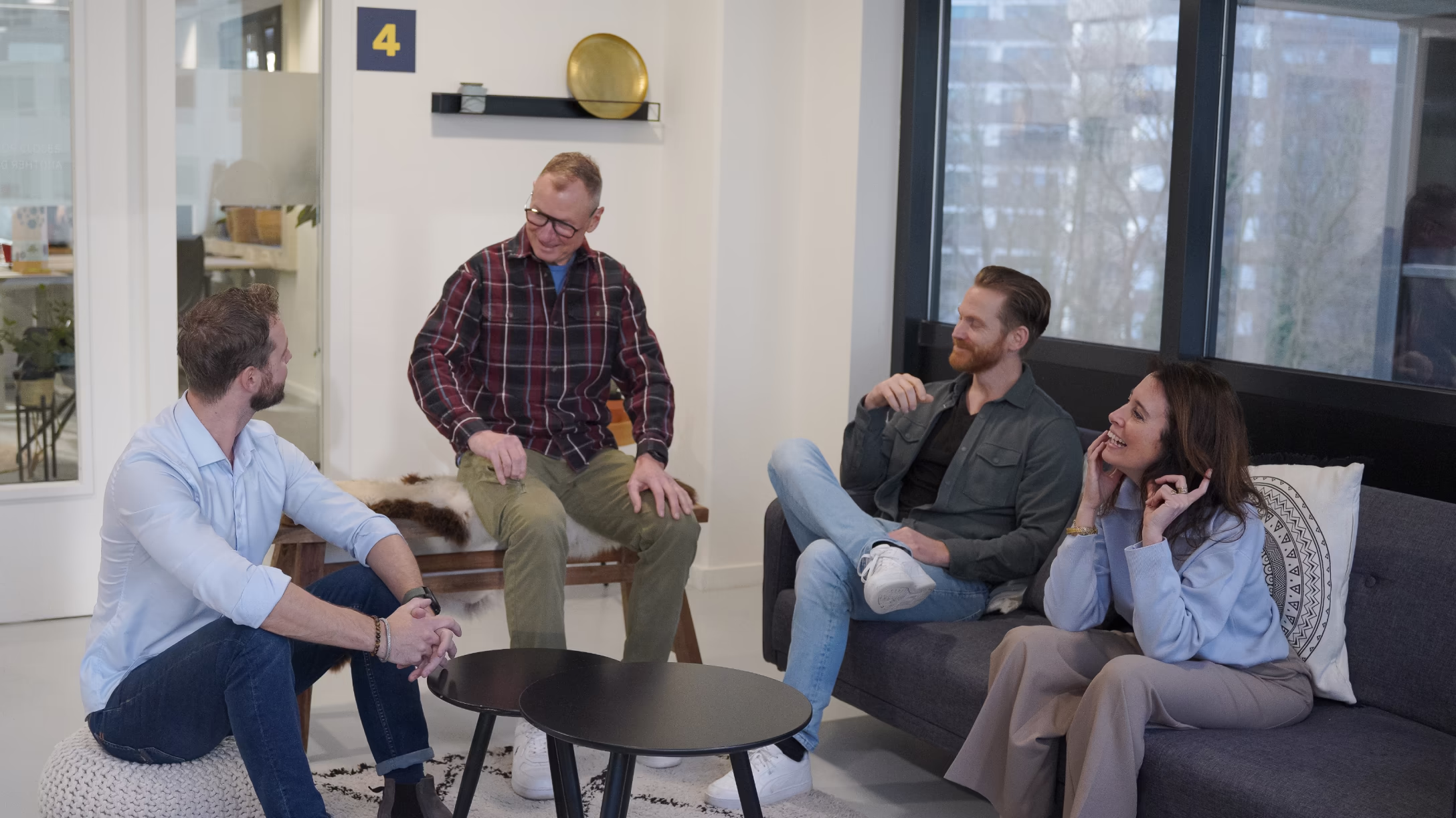 Vier volwassenen zitten in een moderne woonkamerinterieur en voeren een ontspannen gesprek.