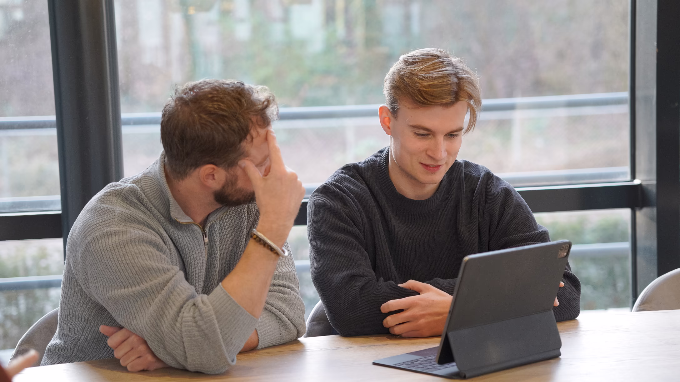 Twee mannen zitten aan een tafel en kijken naar een tablet met toetsenbord in een lichte kamer.