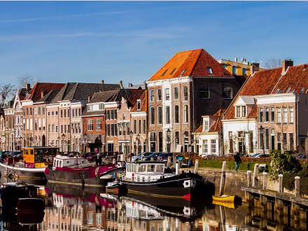 Traditionele Nederlandse huizen langs een rustige gracht met aanlegplaatsen en boten onder een heldere blauwe lucht.