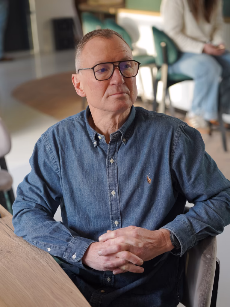 Man met bril en een denim overhemd zit aan een houten tafel met gevouwen handen.