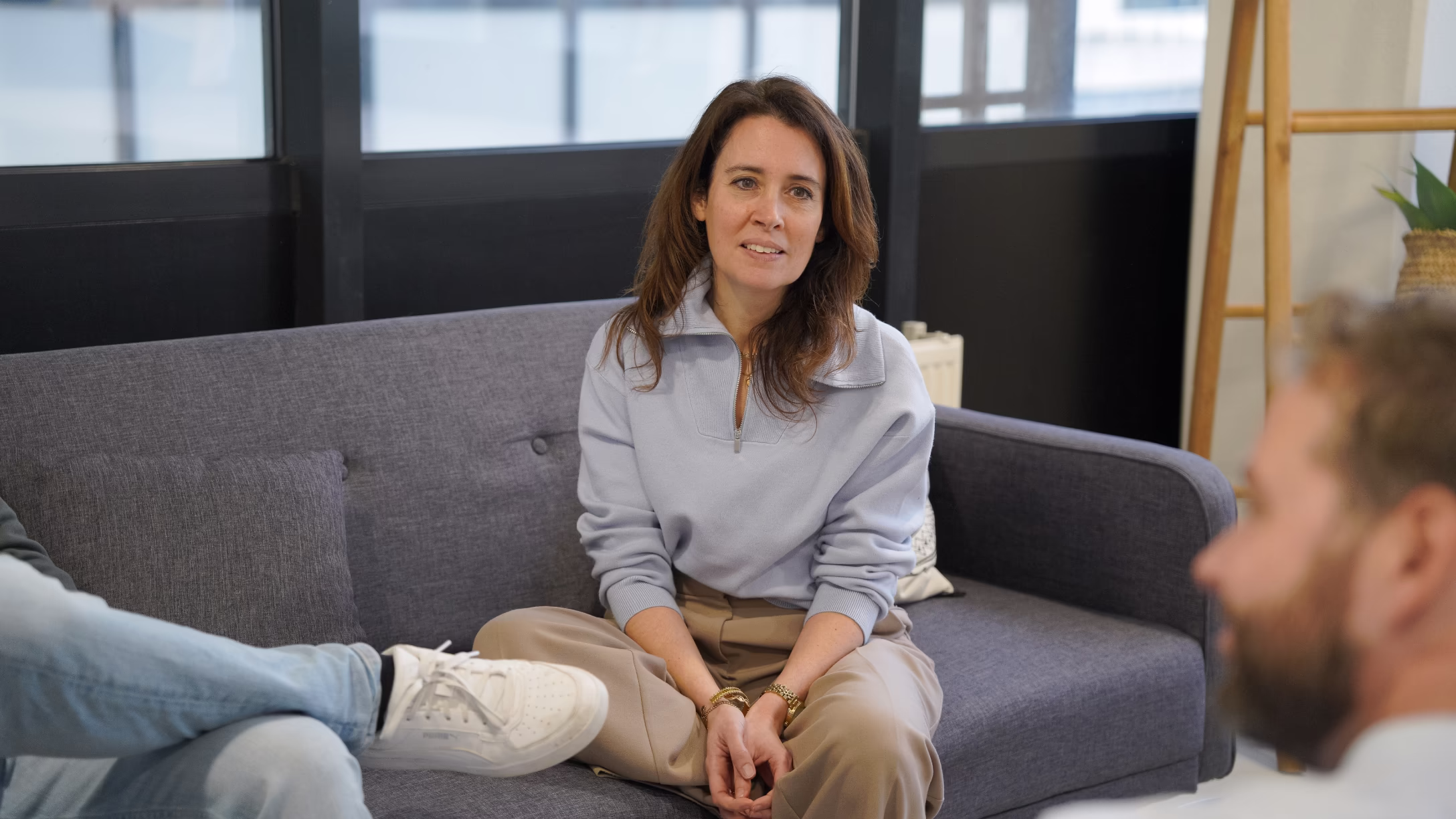 Vrouw zittend op een grijze bank in een kantoorruimte in gesprek met iemand buiten beeld.