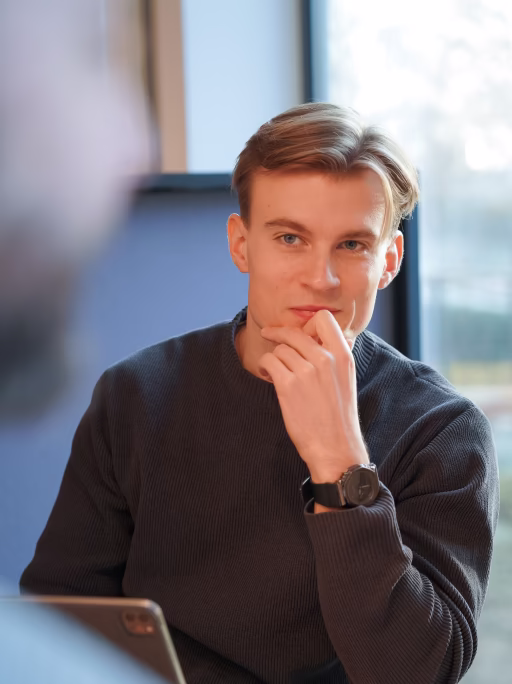 Jonge man met blonde haren en een zwarte trui kijkt bedachtzaam en ondersteunt zijn kin met zijn hand, zittend voor een raam met natuurlijk licht.