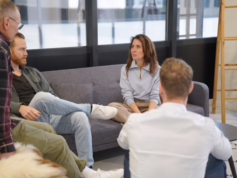 Vier volwassenen zitten in een casual kantoorruimte, terwijl een vrouw op een bank zit en spreekt met de rest van de groep.