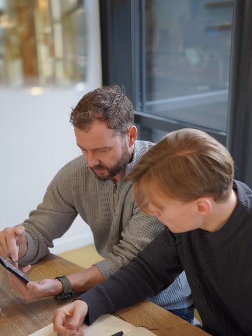 Twee mannen zitten aan een tafel, één toont iets op een smartphone aan de ander.