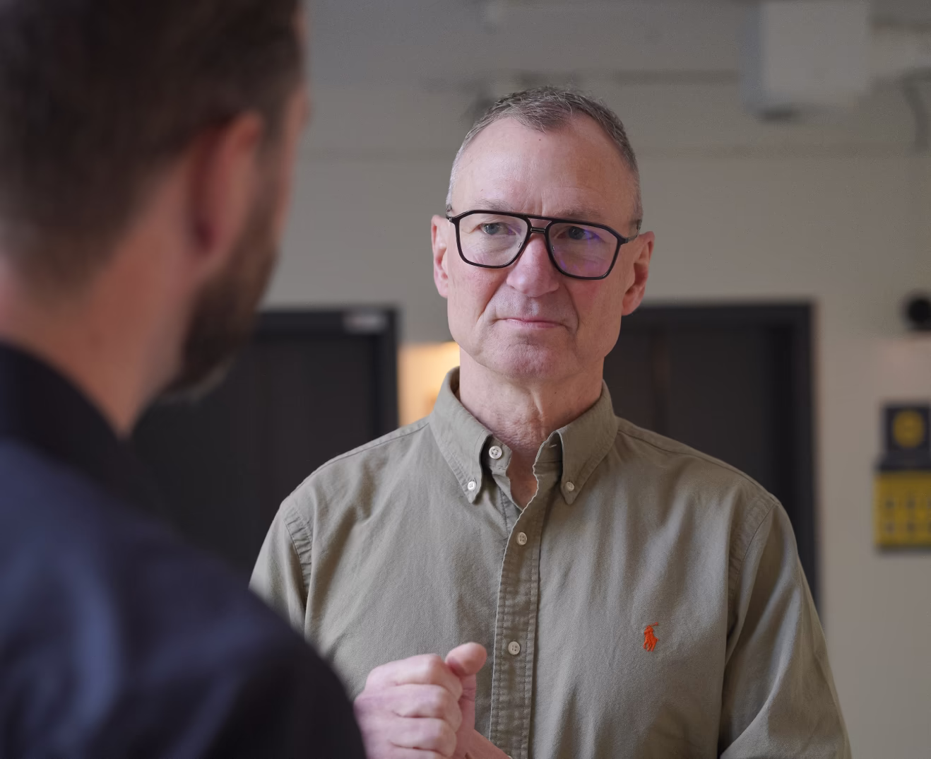 Man met bril en groen overhemd luistert aandachtig tijdens een gesprek met een andere persoon.