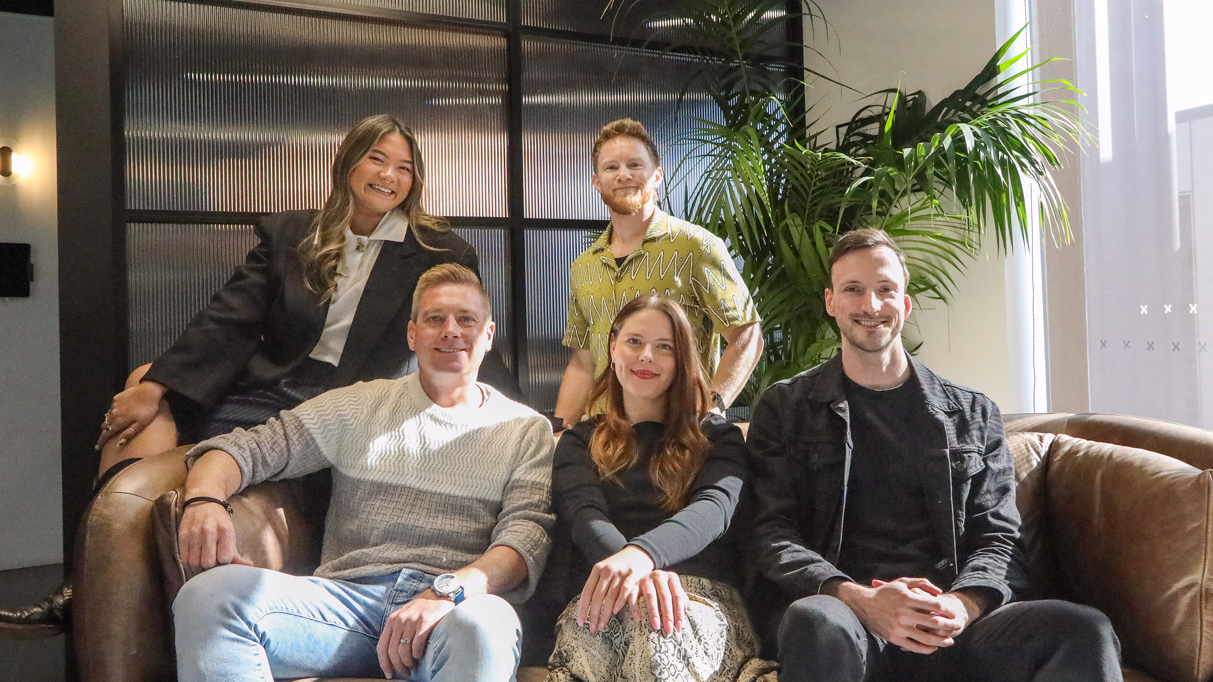 Supersolid team photo in a well lit co-working office. 5 members of the team is gather by a window with a brown leather sofa.
