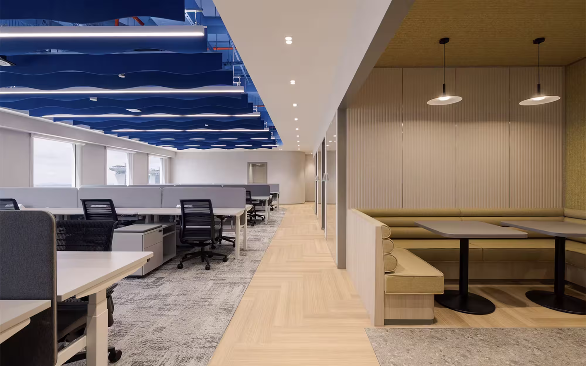 Modern office with rows of desks and black swivel chairs under blue wave-like ceiling panels, with a cosy pendant-lit booth.