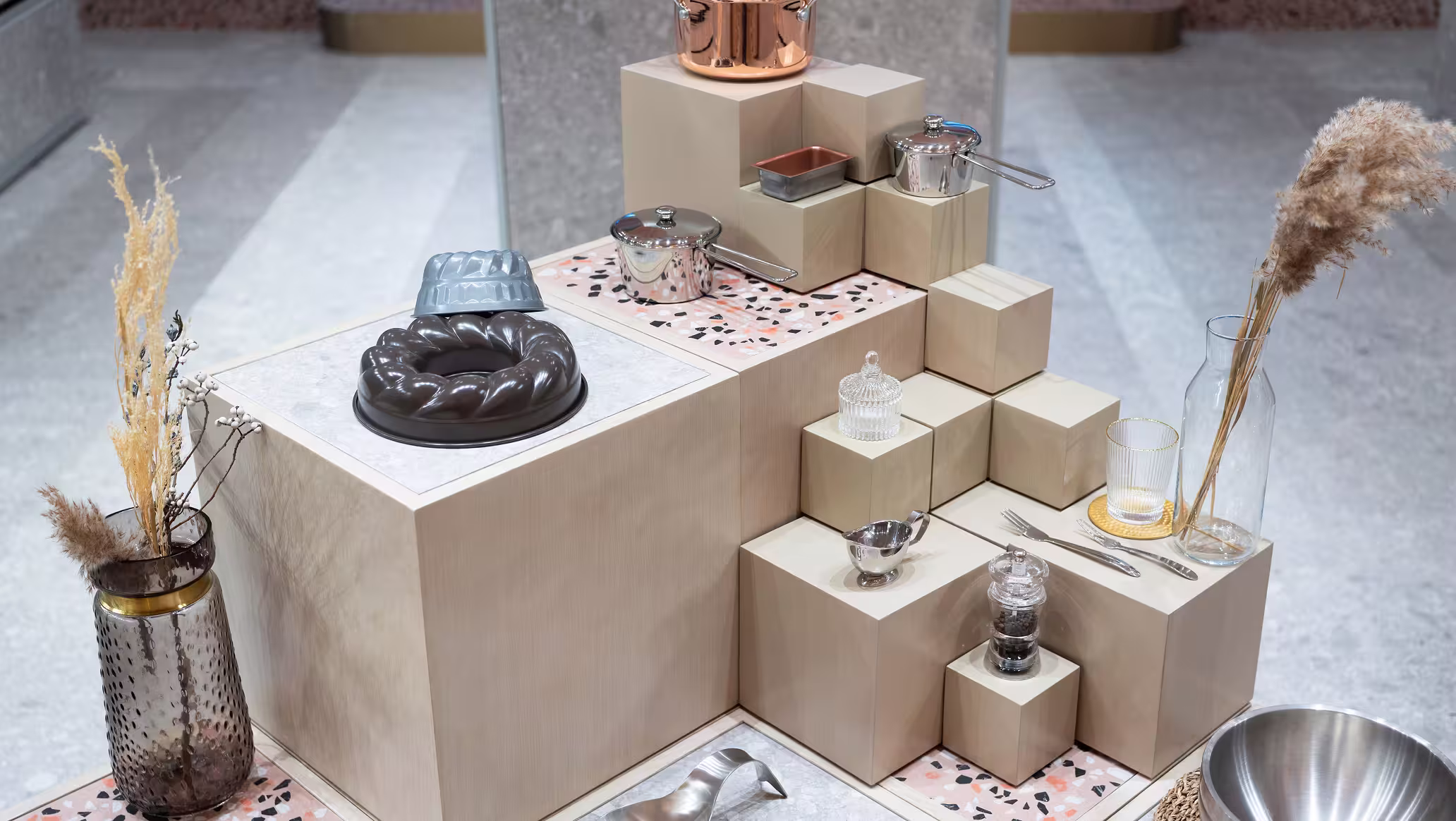 Stacked beige blocks displaying kitchenware including a bundt cake mould, pots, cream pitcher, glass jars, and cutlery.