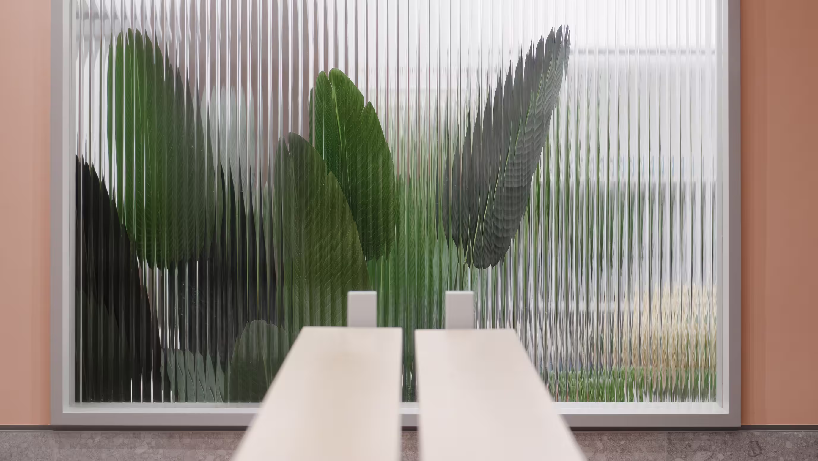Large green tropical leaves visible through a textured glass window with a beige table in front.