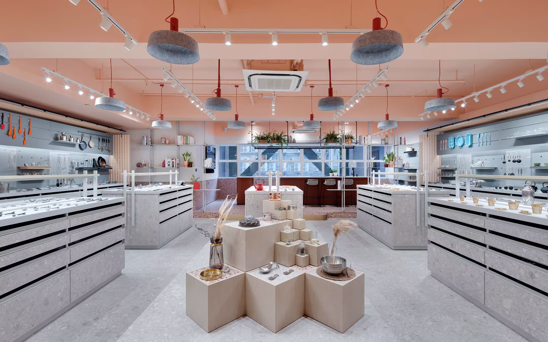 Modern store interior with minimalist display pedestals holding kitchen items, grey cabinets, and large rear windows.