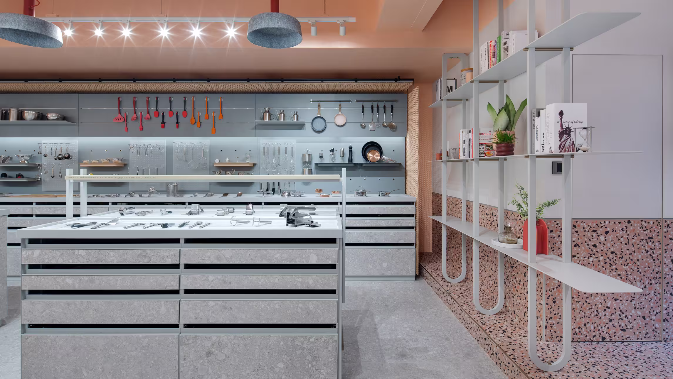 Modern kitchen showroom with utensils displayed on a gray wall and a kitchen island with drawers in the foreground.