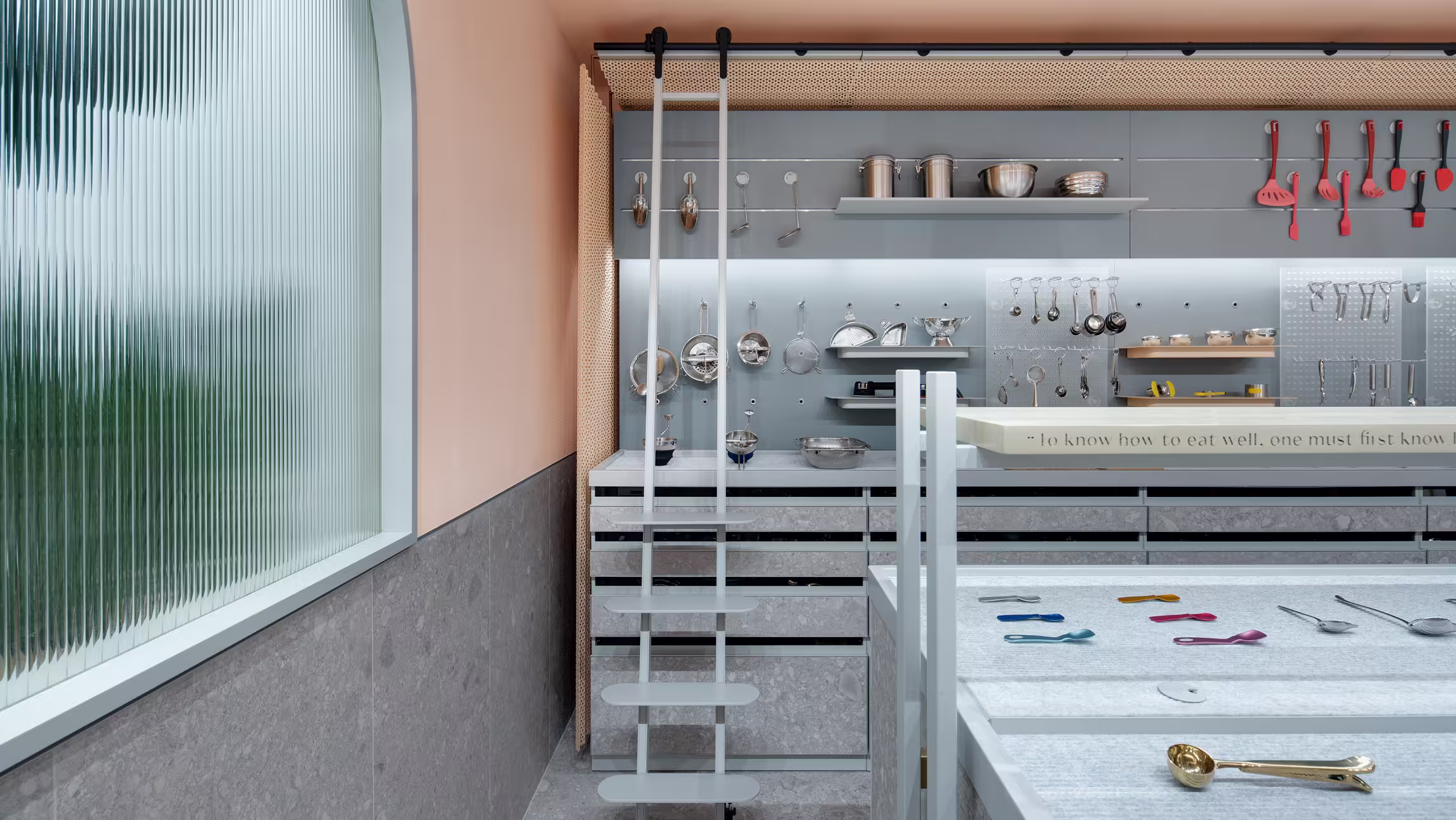 Modern kitchen wall with hanging kitchen utensils, metal shelves, and a sliding ladder beside a large textured glass window.