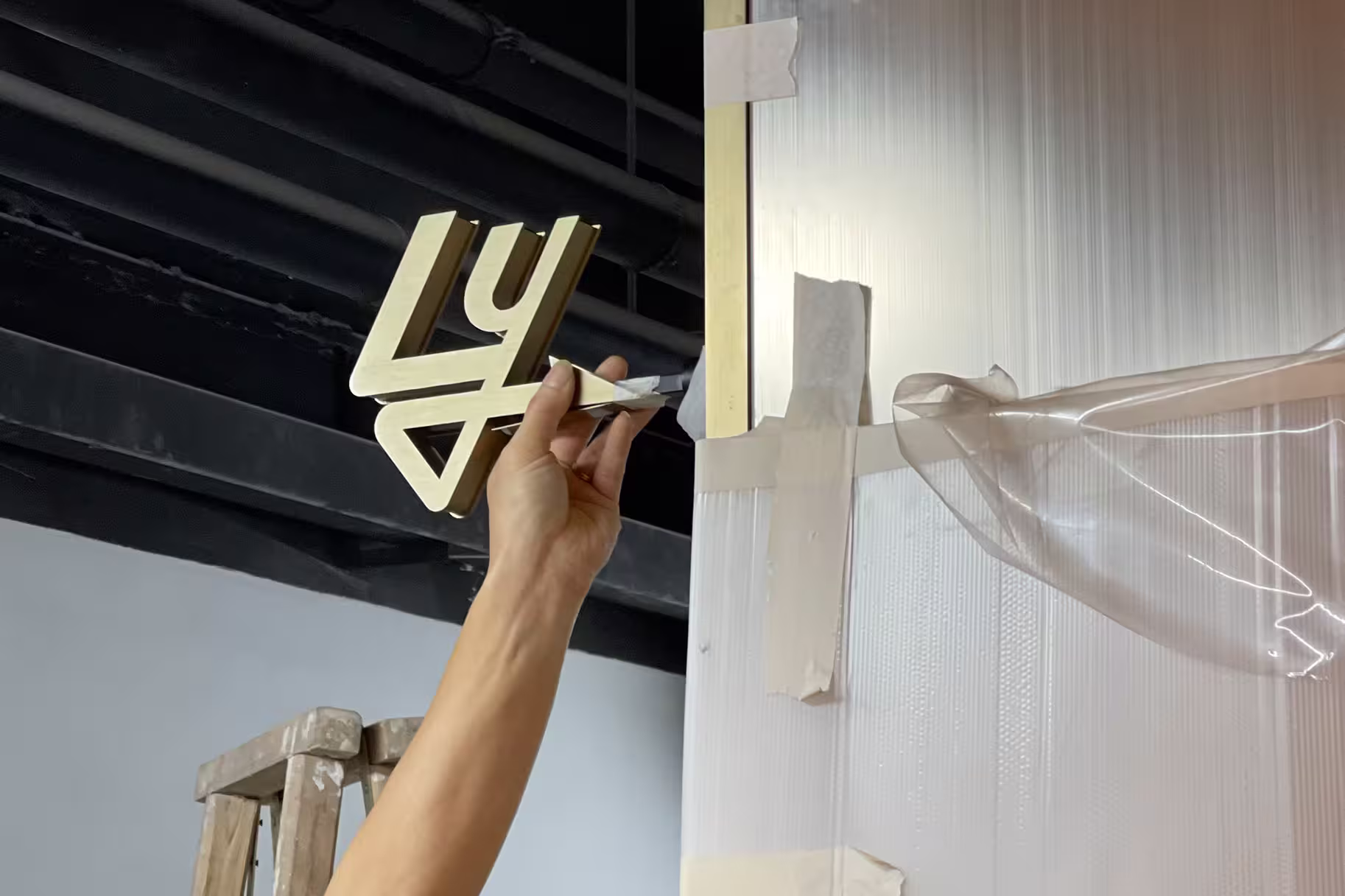 Hand holding a gold decorative number 47 near a taped wall corner with a wooden ladder in the background.