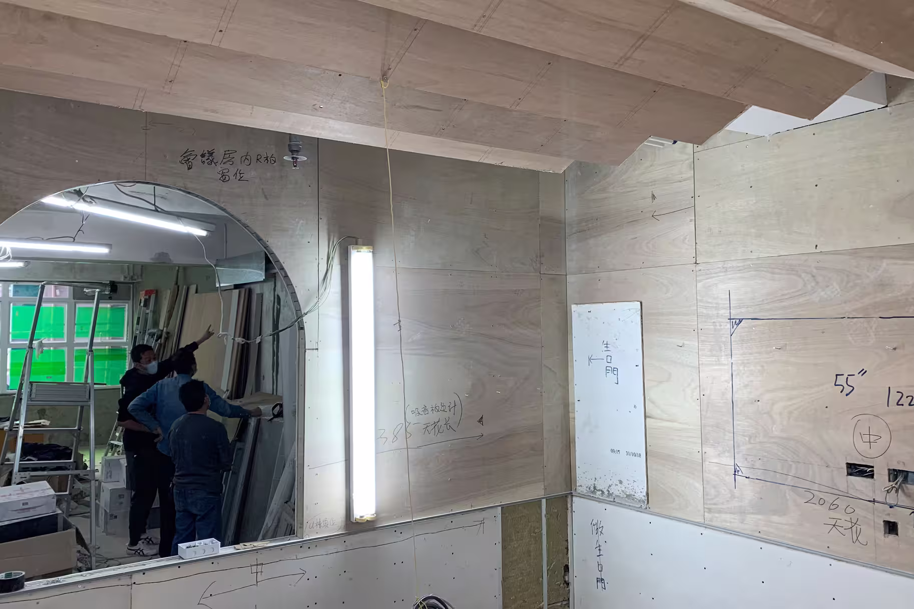 Construction site with three workers discussing plans before an arch-shaped mirror and plywood walls with notes.