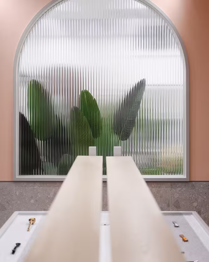 Ribbed glass window framed by a rounded arch and pink walls, with large green leaves blurred behind the glass.
