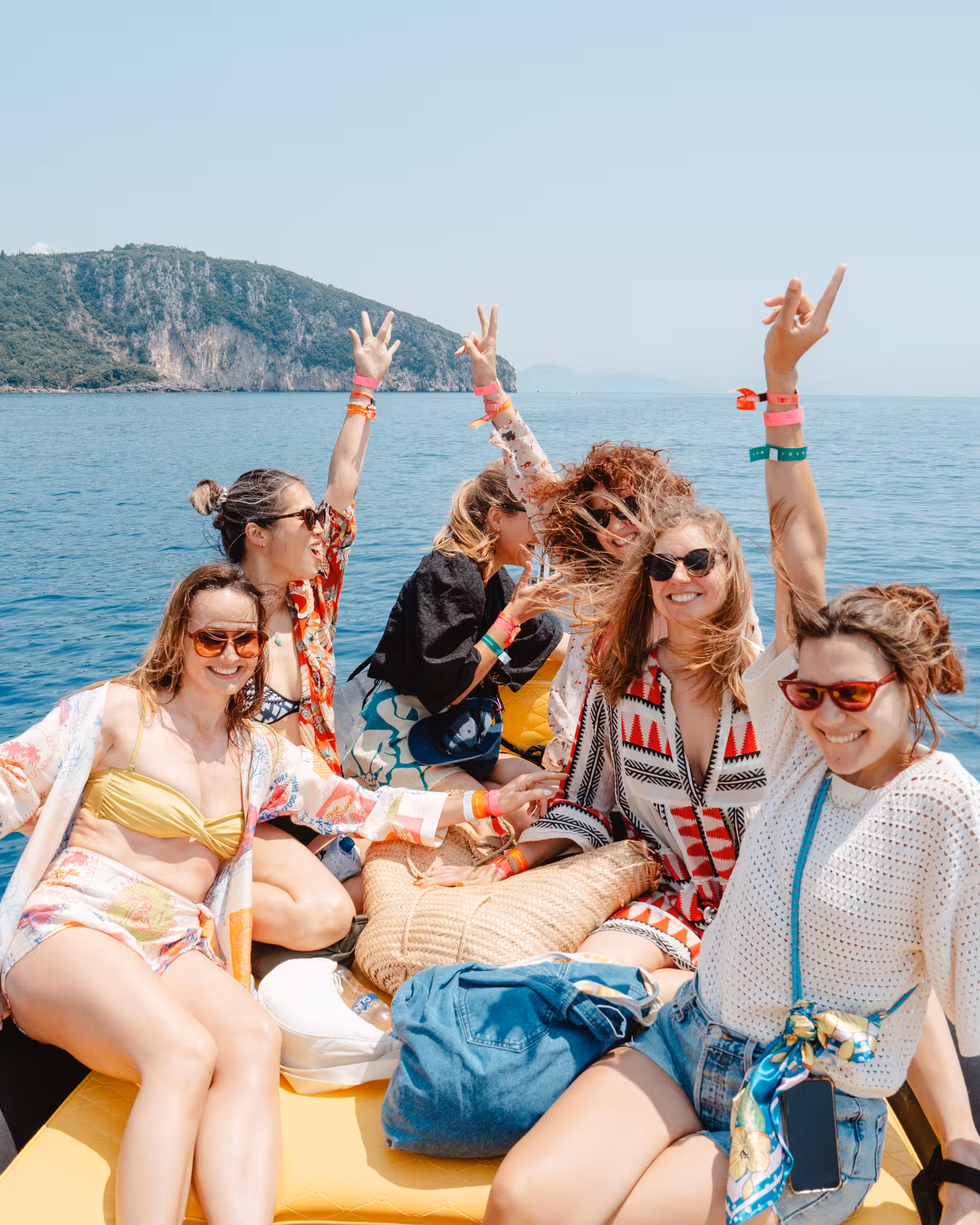 Group of six women smiling and raising their hands while sitting on a boat in open water with a rocky island in the background.