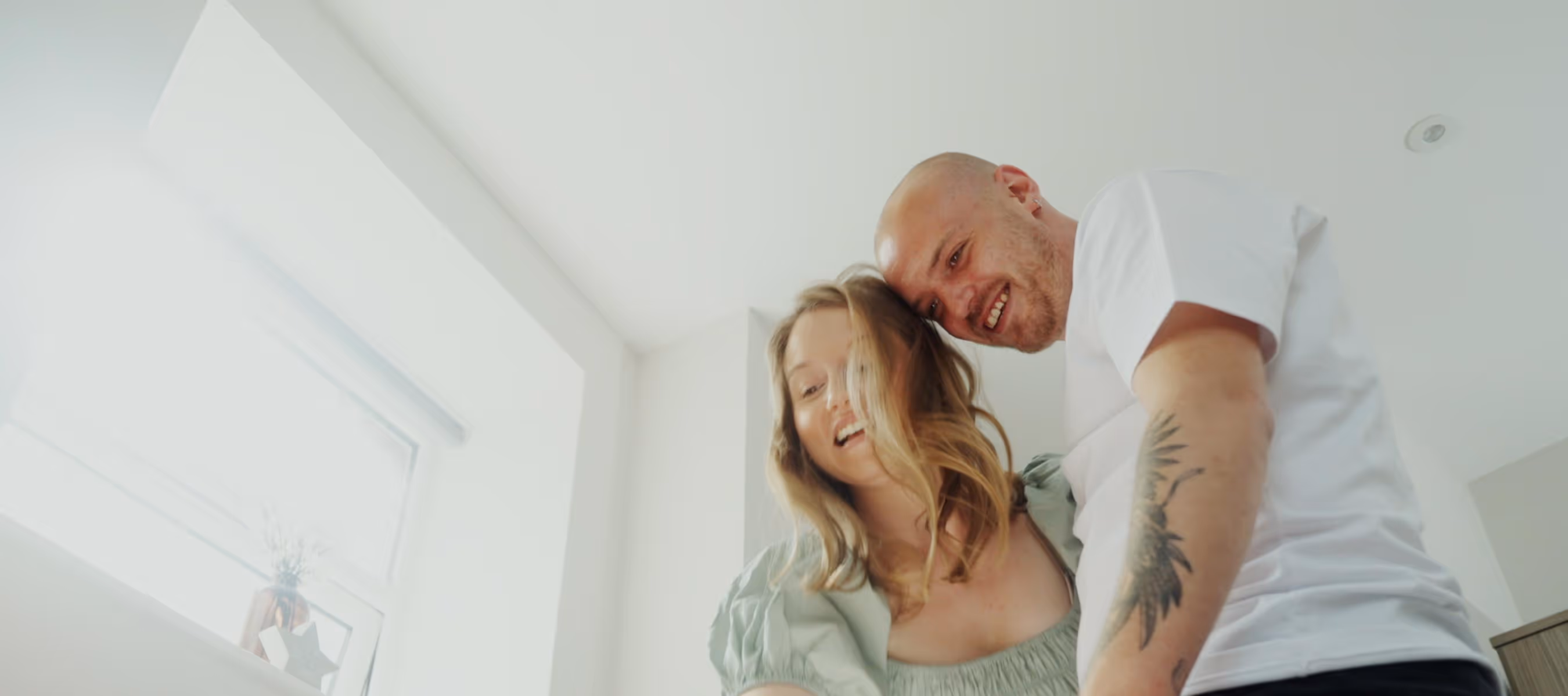 Smiling man with a tattooed arm and woman with wavy blonde hair standing closely in a bright room.