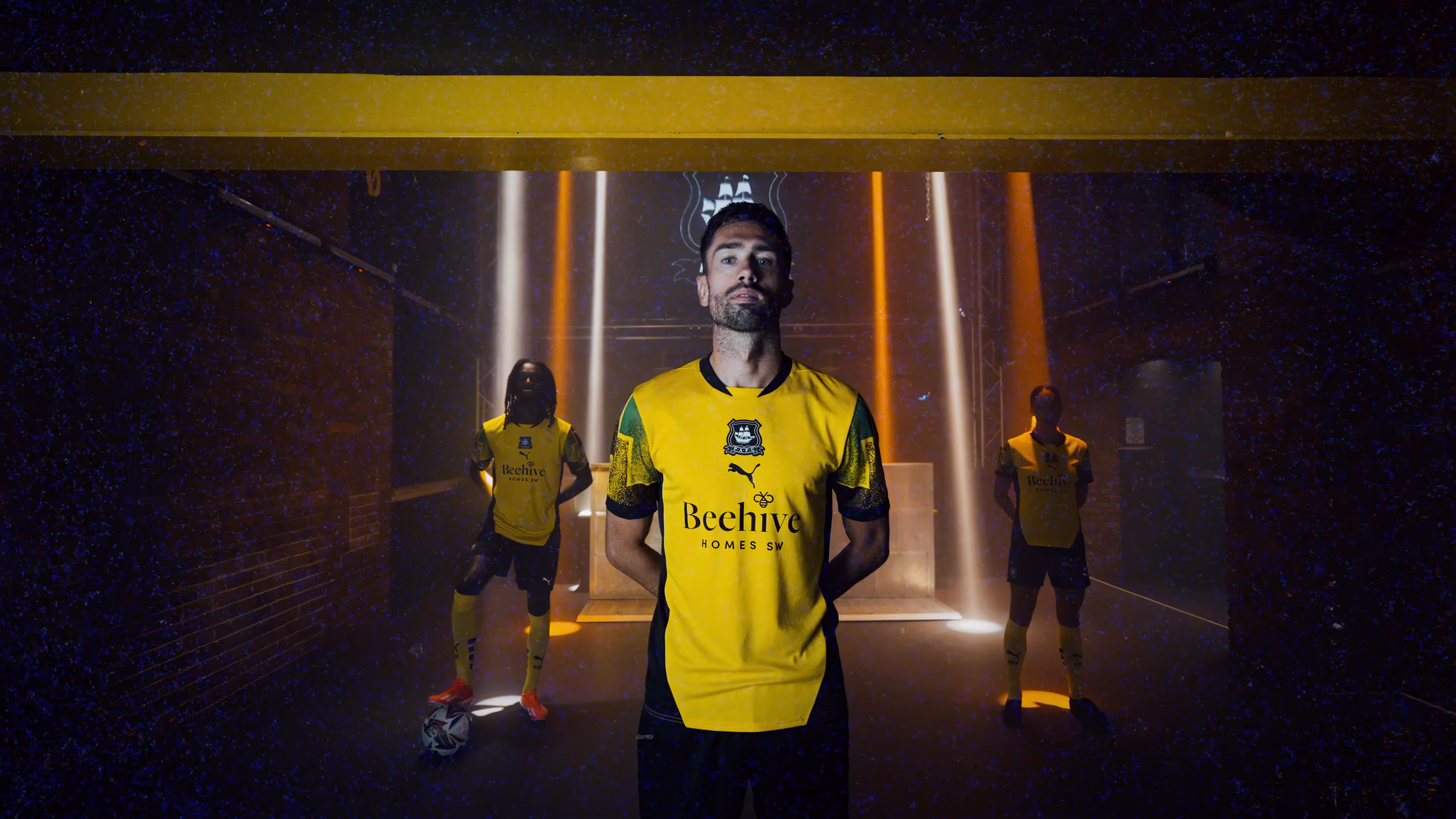 Three soccer players wearing yellow and black uniforms standing under spotlights in a dark indoor setting.
