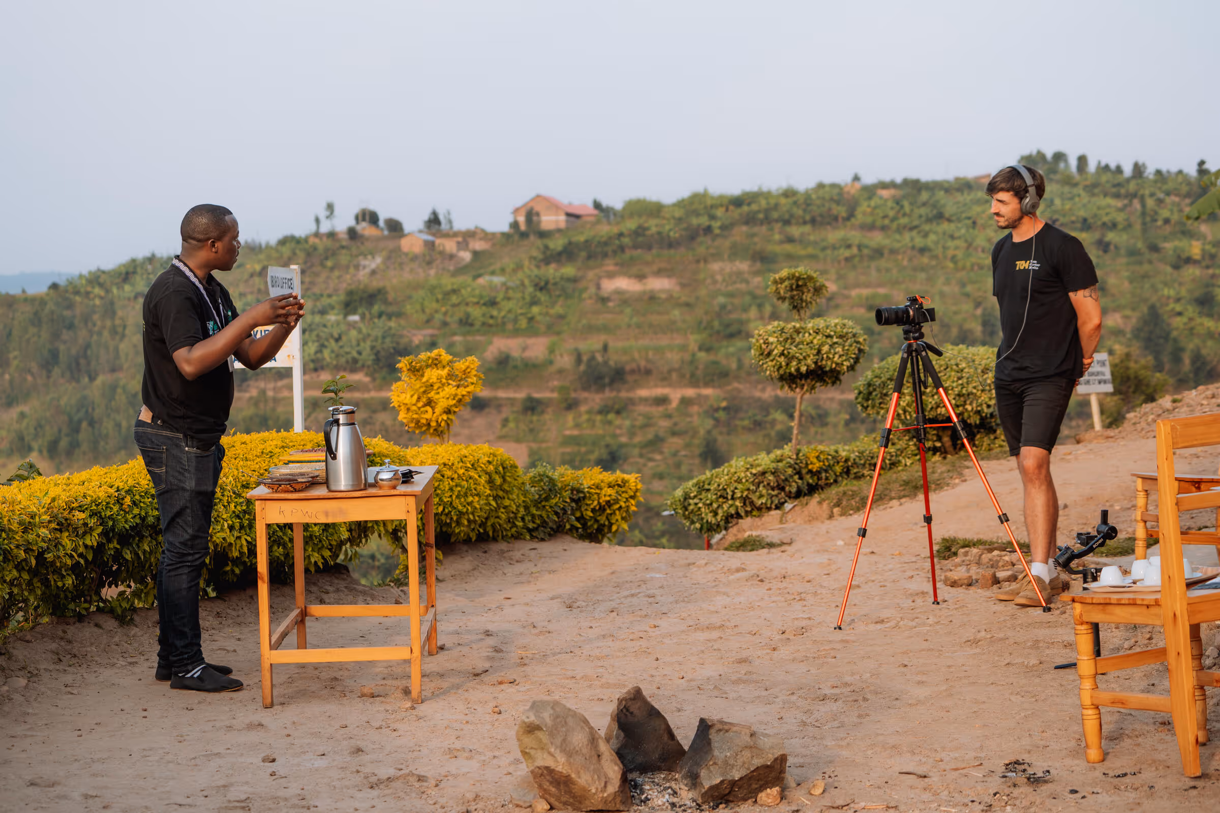Two men outdoors with one standing behind a table holding a cup and the other operating a camera on a tripod, with green hills in the background.