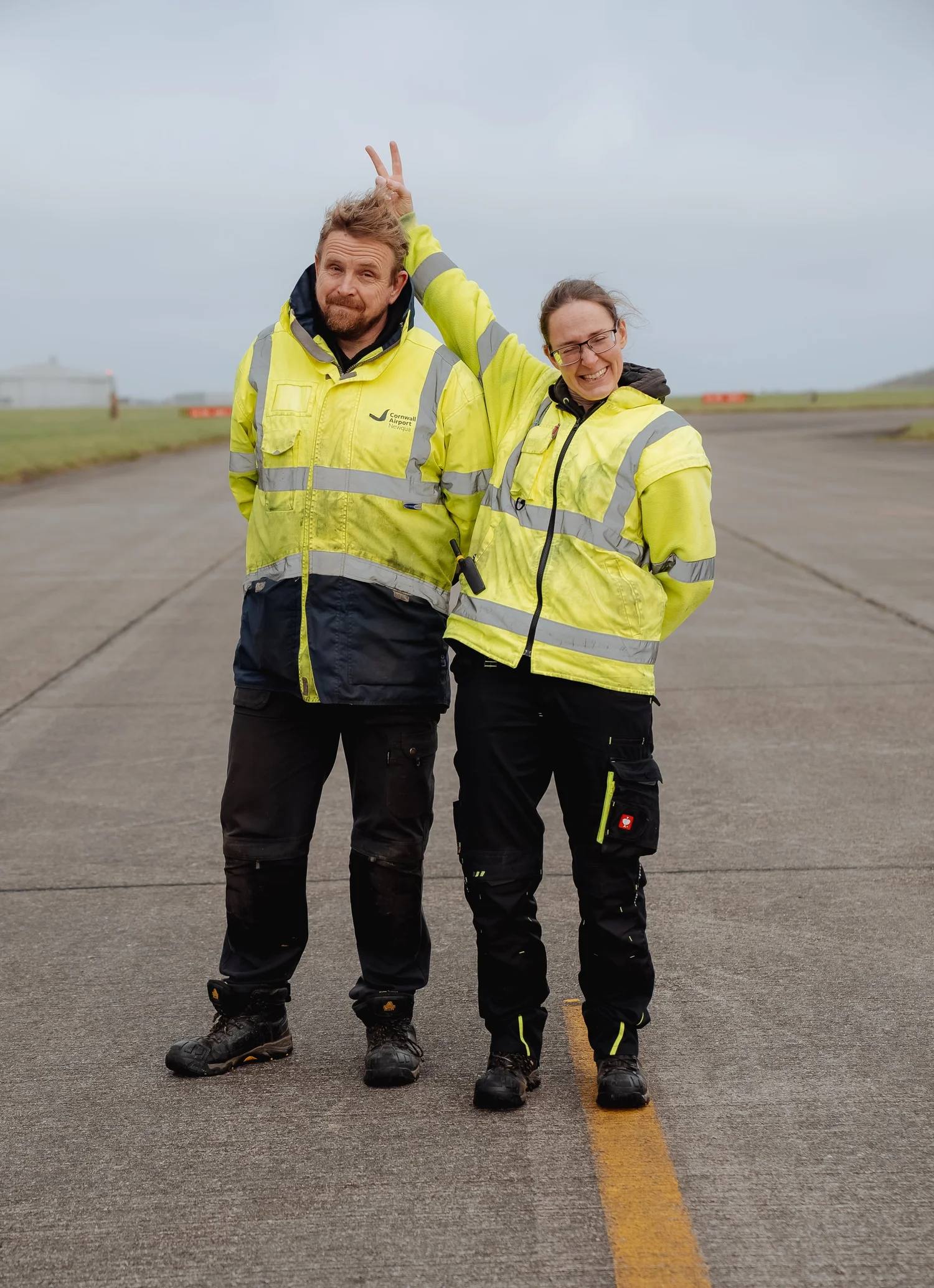 Two people in yellow high-visibility jackets standing on a runway, one playfully making bunny ears behind the other’s head.