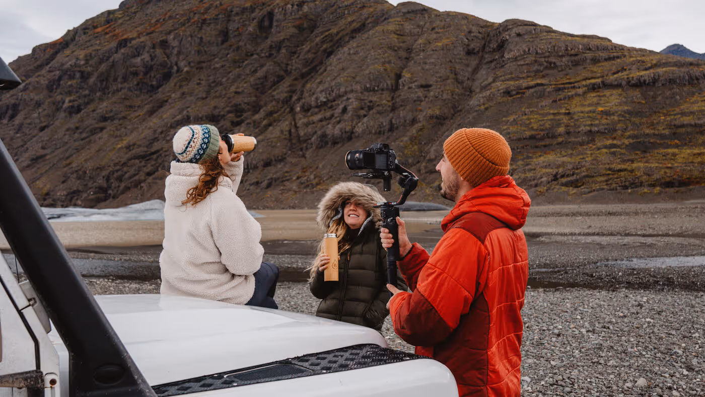 Three people outdoors by a rocky hillside, one woman sitting on a white vehicle drinking from a cup, another woman in a hooded jacket holding a cup, and a man in a red jacket filming with a camera on a stabilizer.