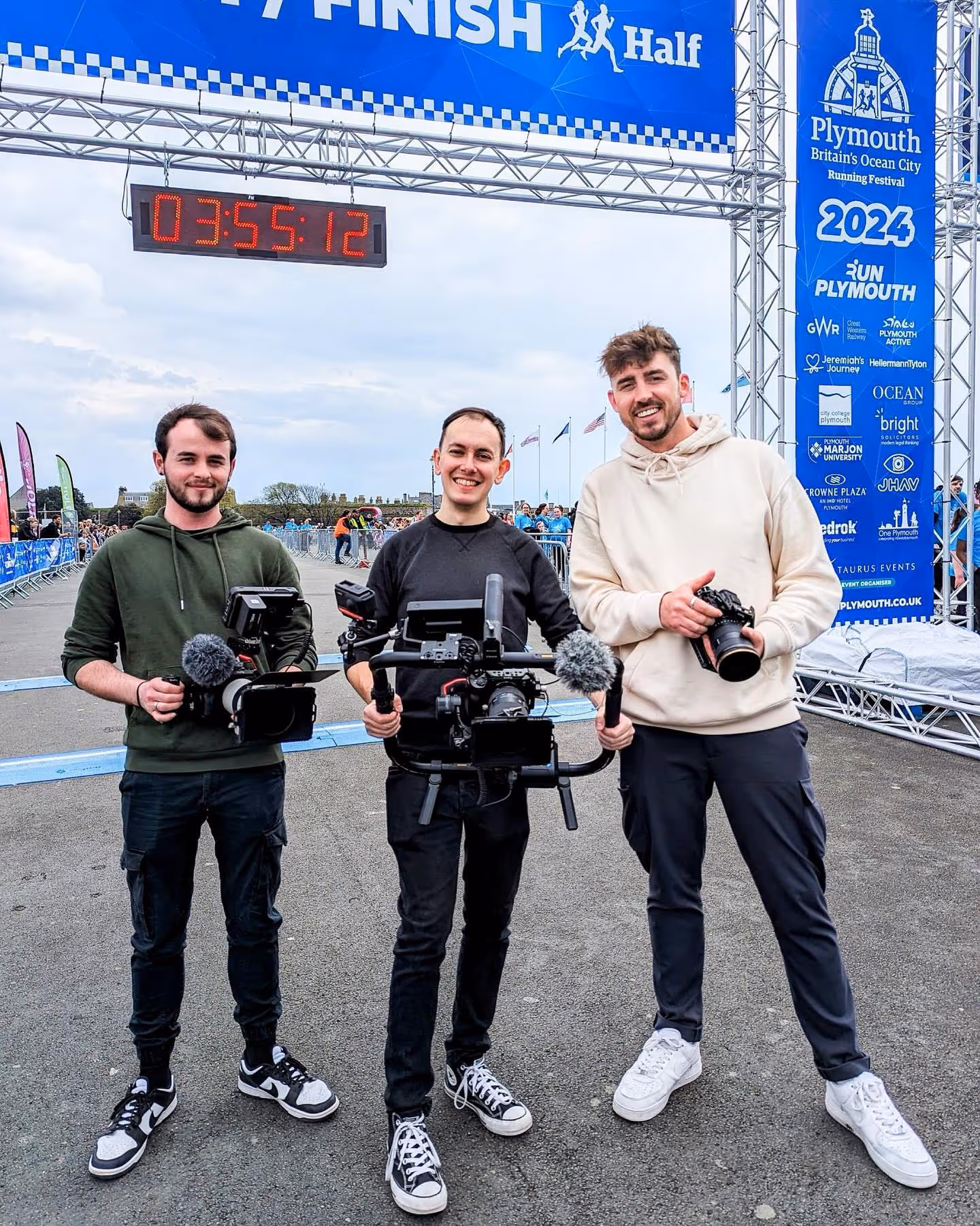 Three men holding professional camera equipment standing under a Plymouth Running Festival finish line banner.
