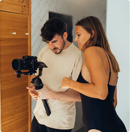 Man holding a camera stabilizer with a camera, showing it to a woman in a black swimsuit indoors.